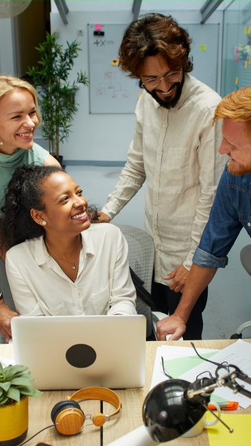 Vertical. Group of professional multirracial men and women working at office. These colleagues look happy. They work around one laptop. Cheerful business team work together at workplace in a meeting